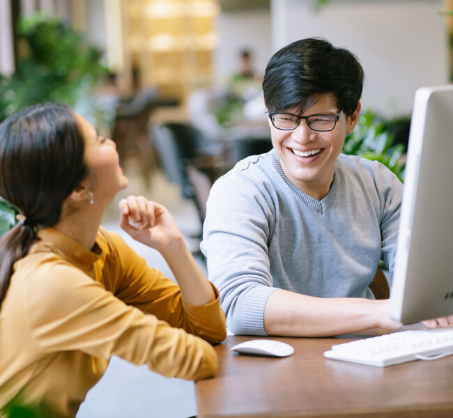 Two,asian,colleagues,sitting,at,desk,talking,relaxed,behind,pc